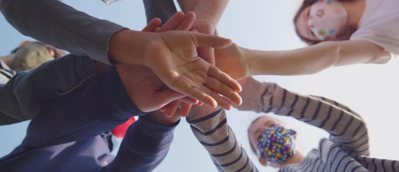 Bottom view of diverse schoolchildren in mask putting hands together outdoors. Preteen classmates in safety mask putting hands together over blue sky background